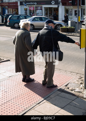 An elderly couple hand in hand waiting to cross the road with the man pushing the crossing button to stop the traffic Stock Photo