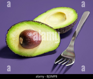 Hass variety avocado pear sliced into two halves along with a fork against a lilac background. Stock Photo