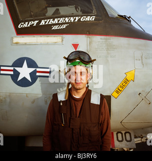 A US Navy electrician looks straight into the camera wearing a brown colour-coded uniform and beneath cockpit of  EA-6B Prowler. Stock Photo