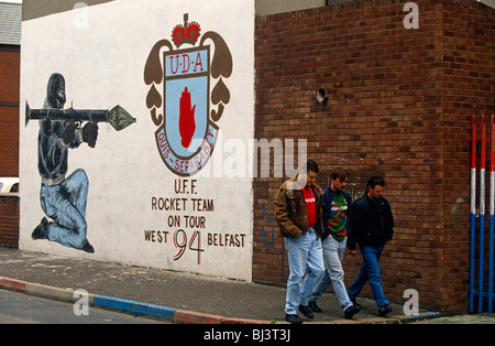 Three men pass along a street off the Shankhill Road in Belfast, under a Loyalist UDA/UFF paramilitary wall mural. Stock Photo
