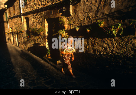 An elderly Spanish lady walks towards strong setting sun that shines through old medieval street in beautiful town Valdemossa. Stock Photo
