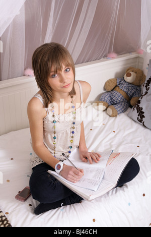 14 year old teenage girl sitting crossed legged on her bed doing homework. Stock Photo