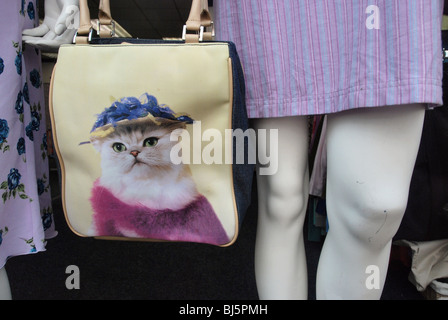 Mannequins in a charity shop window with a kitsch shoulder bag with a design featuring a cat wearing a hat. Stock Photo