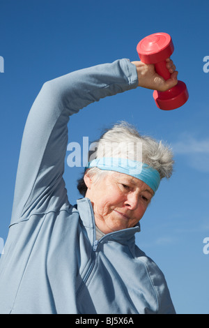 Active senior adult woman wearing a sweatband bending sideways whilst exercising using dumbbell hand weights working out outdoors. England UK Britain Stock Photo
