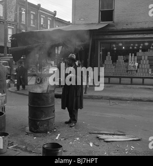 A well dressed African-American man warms his hand over a barrel fire in Philadelphia during the early 1950s. storefront poor Stock Photo