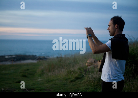 man photographing the sea from grassy hill Scotland near Edinburgh on a warm summer evening on a camping trip Stock Photo