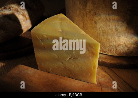 Wedge of mature cheddar cheese on board with whole cheese and bread in background Stock Photo