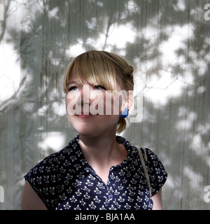 portrait of young blonde woman with fringe and patterned dress posing infront of white wall covered in shadow of leaves Stock Photo
