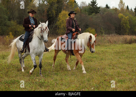 Two women dressed in cowboy clothes and hats riding horses western style on horseback at a ranch Stock Photo