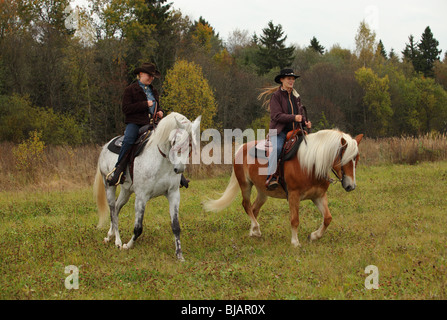 Two women dressed in cowboy clothes and hats riding horses western style on horseback at a ranch Stock Photo