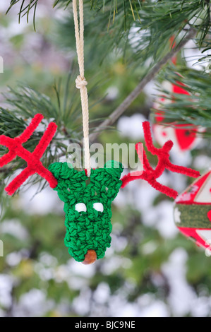 Funny green and red handmade reindeer Christmas ornament hangs from snowy bough of pine tree (pinaceae) (pinus nigra) copy space Stock Photo