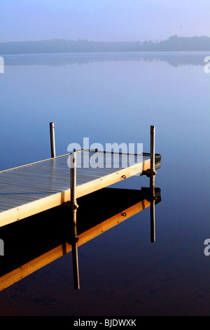 A lonely pier in early morning mist reflects a mirrored image in northern WI lake creates a magical view of the distant horizon Stock Photo
