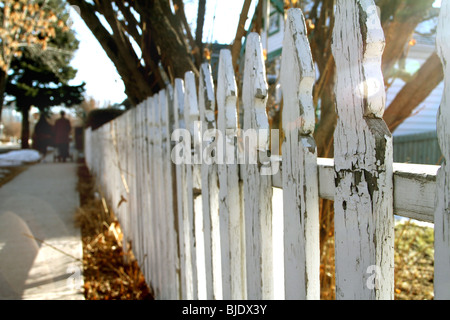 Sun shines through a white picket fence as a couple walk down the street with their child in a stroller. Stock Photo