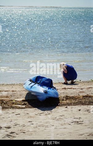 WOMAN KNEELING ON SHORE NEAR KAYAK COLLECTING SEA SHELLS Stock Photo