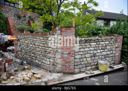 Building a garden wall with cotswold stone and cement mortar with reclaimed red brick pillars and coping UK Stock Photo