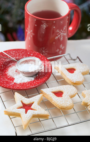 Jam biscuits on cake rack, icing sugar, mug of tea - Stock Photo