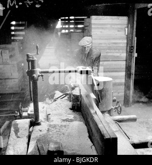 England,  1950s. Male construction worker, wearing cap and jacket, operating a wood cutting machine. Stock Photo