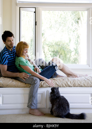 couple sitting on a window bench petting a cat Stock Photo