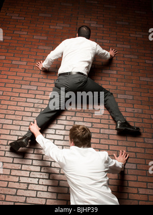 man climbing up a brick wall while another man grabs on to his leg Stock Photo