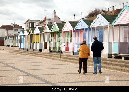 Two people walking past beach huts on the promenade, Southwold, Suffolk, UK