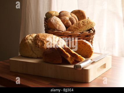 Selection of fresh bread on a wooden chopping board, includes a basket of assorted rolls in a wicker basket in the background. Stock Photo