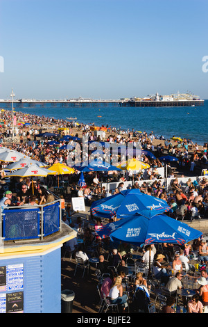 England, East Sussex, Brighton, People on beach and at bar tables on seafront promenade with Pier beyond Stock Photo