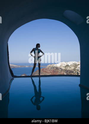 Silhouette of woman in bikini standing at edge of infinity pool with arch and rock formation Stock Photo