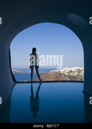 Silhouette rear view of woman in bikini standing at edge of infinity pool with arch and rock formation Stock Photo