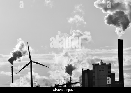 Black-and-white image of smoke billowing into the sky from a factory's smokestacks as a wind turbine stands in the foreground Stock Photo