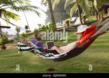 Man wearing a hat, lying in a hammock under palm trees and relaxing while working with a netbook, Bethsaida Hermitage near Kova Stock Photo