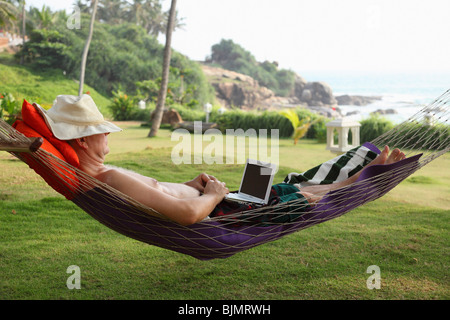Man wearing a hat, lying in a hammock and relaxing while working with a netbook, Bethsaida Hermitage near Kovalam, Kerala, sout Stock Photo