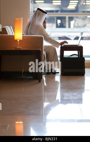 An anonymous Arab man in traditional clothing of Thoub or dish dash and shemagh using a laptop computer inside a modern building Stock Photo
