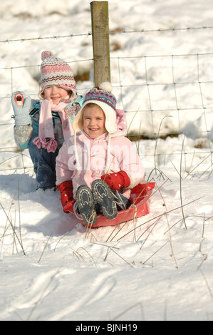 Two girls in ski coats and woolen hats having fun tobogganing in the snow with a red sledge Stock Photo