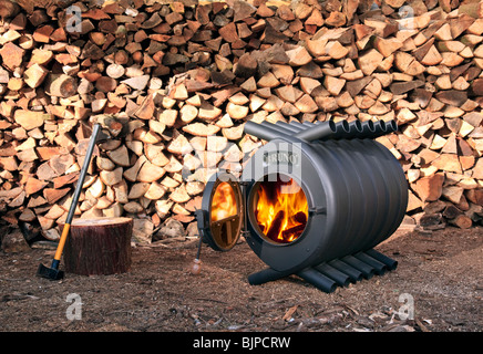 A large wood burning stove in front of  a pile of logs, with an axe nearby Stock Photo