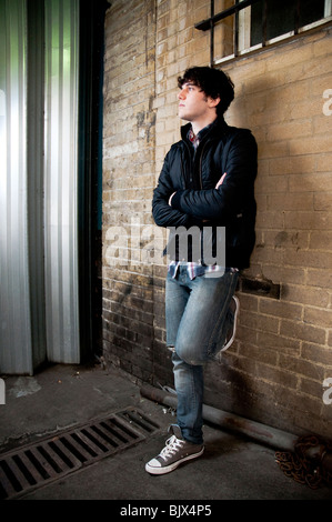 Full body street shot of young man leaning against brick wall looking away. Stock Photo