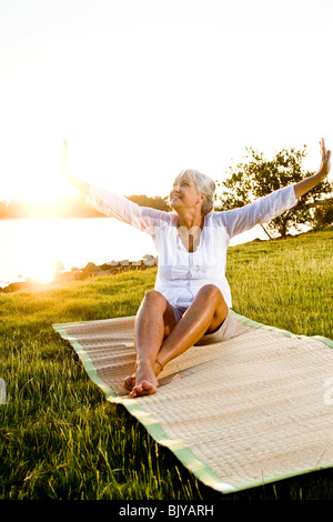 Middle-aged woman smiling and stretching outdoors on grassy shoreline Stock Photo