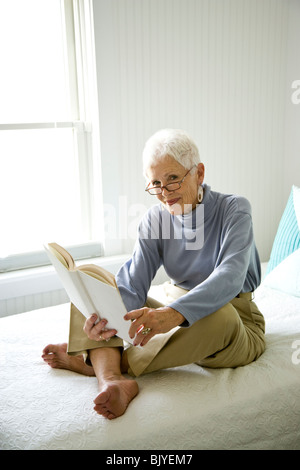 Portrait of senior woman sitting on bed with book smiling Stock Photo
