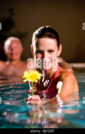Middle-aged woman holding flower in swimming pool, man in background Stock Photo