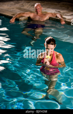 Middle-aged woman holding flower in swimming pool, man in background Stock Photo