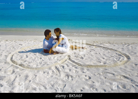 Young couple on beach sitting in a heart shaped imprint on the sand, Maldives, Indian Ocean, Asia Stock Photo