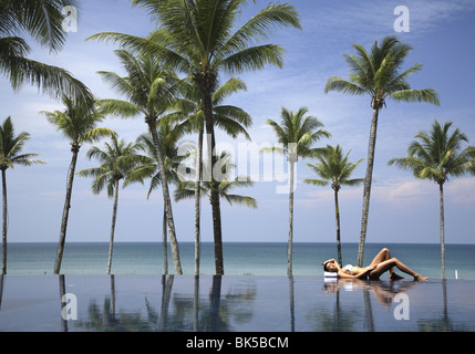Young woman relaxing by pool with palm trees and the sea beyond Stock Photo