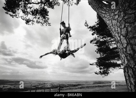 A couple of trapeze artists practice their act in the open air, swinging from the branch of a big tree Stock Photo