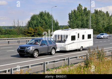 Land Rover car towing white caravan side front view driving along  rural section of M11 UK motorway road steel crash barriers Essex England UK Stock Photo