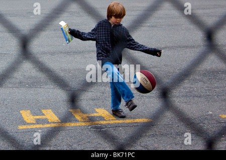 Boy kicking ball, holding book (The Secret Garden) on a school playground, Manhattan, New York City Stock Photo