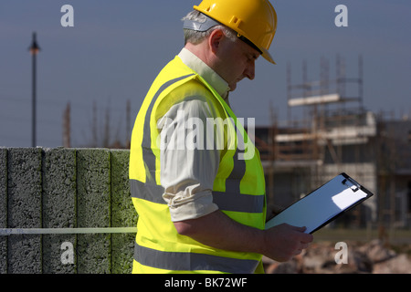 middle aged grey haired man wearing hard hat and hivis vest holding and looking down at a clipboard building blocks Stock Photo
