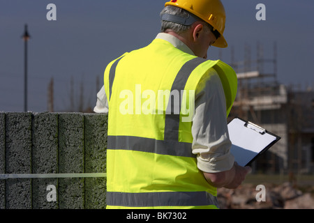 middle aged grey haired man wearing hard hat and hivis vest holding and looking down at a clipboard reviewing plans checking specs building blocks Stock Photo