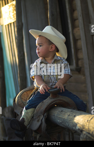 A cute 2-4 year old mixed race boy sitting on a saddle wearing a cowboy hat.(Released) Stock Photo