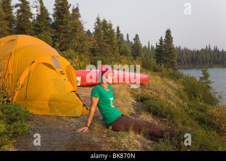 Young woman sitting, enjoying view, relaxing, tent and canoe behind, Caribou Lakes, upper Liard River, Yukon Territory, Canada Stock Photo
