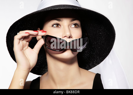 Young woman wearing a black hat and a black dress holding sunglasses in front of her face Stock Photo