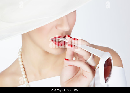 Portrait of a young woman with a white summer hat, a white dress and a pearl necklace with the arm of a pair of white sunglasse Stock Photo